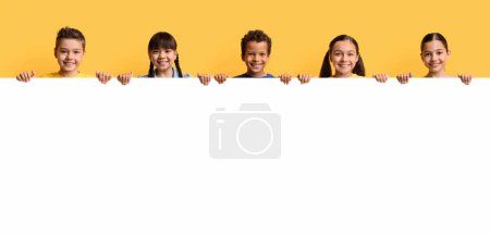 Téléchargez les photos : Joyeux groupe souriant d'enfants multiraciaux, d'amis, de garçons et de filles, montrant la maquette horizontale vierge de tableau blanc vide pour le texte, la publicité sur fond jaune, la bannière - en image libre de droit