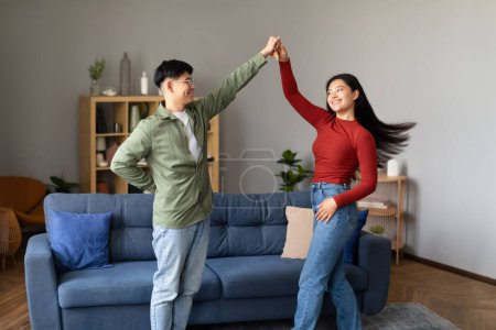 Téléchargez les photos : Heureux japonais amoureux couple dans casual danse avoir amusant à la maison. Jeune mari et femme qui aiment danser en se tenant la main dans le salon intérieur confortable. Fête domestique, date romantique - en image libre de droit
