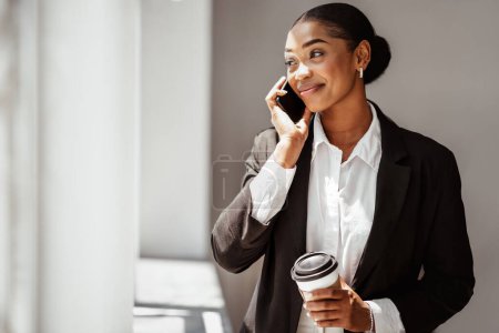 Photo for Happy attractive black lady entrepreneur in smart casual drinking takeaway coffee holding paper cup and talking on phone in office interior, copy space - Royalty Free Image
