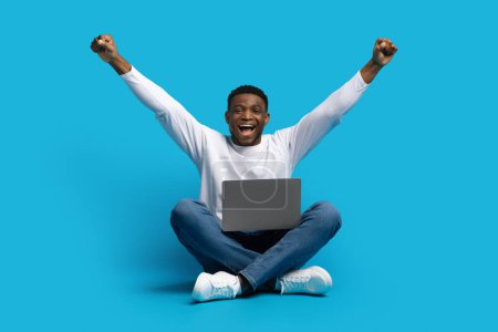 Foto de Emocional joven negro feliz sentado en el suelo, usando computadora portátil, levantando las manos y exclamando, suerte afroamericano chico jugando o comerciando en línea, aislado en fondo azul - Imagen libre de derechos