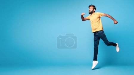 Foto de Divertido hombre indio de mediana edad corriendo sobre fondo de estudio azul, vista lateral de alegre macho saltando en el aire, darse prisa a las ventas o descuentos, panorama, espacio de copia - Imagen libre de derechos