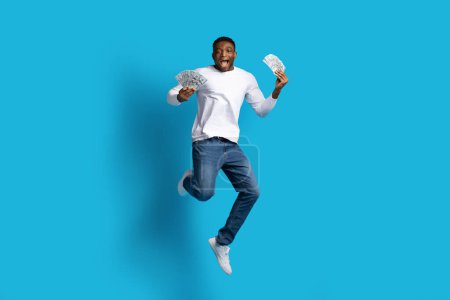 Photo for Cashback, loan, giveaway. Rich wealthy excited handsome young black guy in casual outfit jumping in the air with money cash dollar banknotes in his hands over blue studio background, copy space - Royalty Free Image