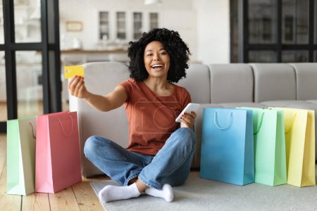 Foto de Concepto de cibercompra. Mujer afroamericana feliz con teléfono inteligente y tarjeta de crédito sentado en el suelo entre las bolsas de compras brillantes, dama negra pidiendo cosas en línea, recomendando compras por Internet - Imagen libre de derechos