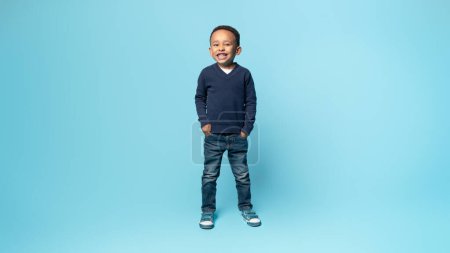 Photo for Full length shot of glad african american little boy posing holding hands in pockets, standing on blue studio background and smiling at camera, panorama, free space - Royalty Free Image