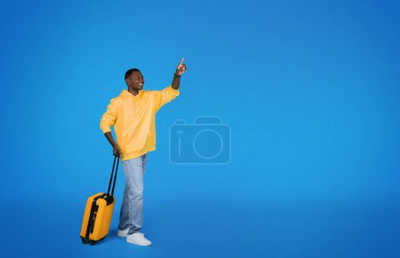 Photo for Cheerful millennial african american guy tourist in sweatshirt with suitcase point finger on free space, isolated on blue studio background. Ad and offer, adventure, travel and tourism - Royalty Free Image