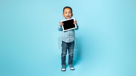 Photo for Cute african american little kid boy in casual holding horizontal digital tablet with black blank screen, isolated on blue background, showing nice app for kids, mockup, copy space - Royalty Free Image