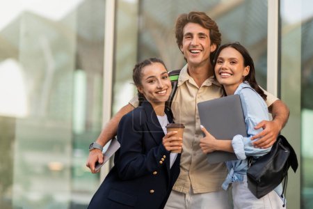 Photo for Happy cheerful handsome guy and two pretty ladies students with books, laptop in hands embracing and smiling outdoor, friends having fun after school, drink takeaway coffee, walk by city, copy space - Royalty Free Image