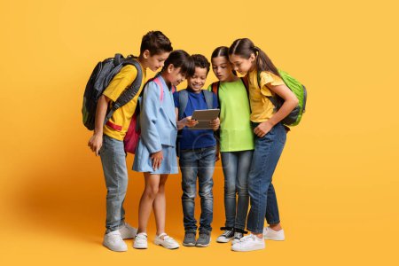 Photo for Pupils, Technology And Learning Concept. Smiling cute boy holding digital tablet, showing group of multiracial classmates friends nice education app, studying at elementary school, yellow background - Royalty Free Image