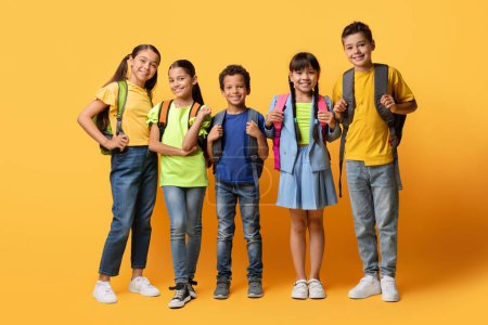 Foto de Concepto de Educación. Grupo de escolares diversos niños y niñas felices posando mirando a la cámara, vistiendo ropa casual y llevando mochila, fondo de estudio de color naranja amarillo, pancarta - Imagen libre de derechos