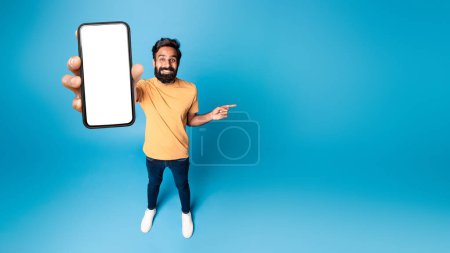 Photo for Mobile offer. Happy indian man showing phone with blank screen and pointing finger aside at free space on blue studio background, panorama, mockup. Choose this application - Royalty Free Image