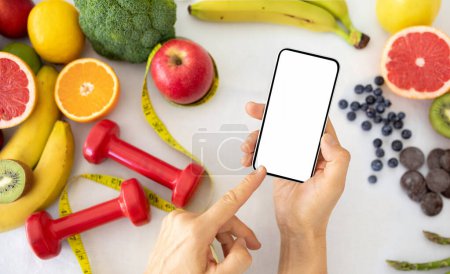 Téléchargez les photos : Main de nutritionniste caucasien montre un plan de perte de poids au téléphone avec écran blanc sur la table avec haltères, légumes et fruits biologiques. Application de soins de santé, bonne nutrition, gadget - en image libre de droit