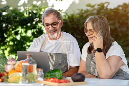 Photo for Smiling old caucasian husband and wife in aprons use laptop, make phone calls at table with organic vegetables in home garden, outdoor. New recipe to cook food, device for remote communication - Royalty Free Image