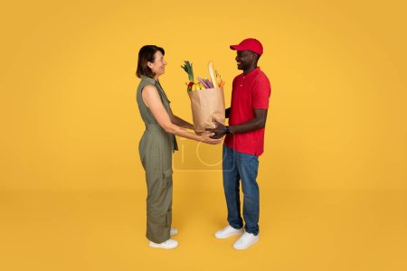 Photo for Glad millennial african american man courier in uniform give paper bag with food to adult woman, isolated on orange studio background, full length. Delivery at home, work and service - Royalty Free Image