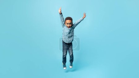 Téléchargez les photos : Petit garçon africain américain ludique sautant les mains en l'air, s'amusant et souriant, fond bleu, pleine longueur, panorama, espace libre. Loisirs d'enfance, concept de bonheur - en image libre de droit