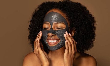 Photo for Closeup of smiling attractive millennial topless african american lady using black mask, isolated on brown background. Peel-off face mask for deep skin cleanse, face care routine concept - Royalty Free Image