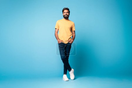 Photo for Positive handsome middle aged indian man wearing casual outfit posing with hands in pockets on blue studio background, smiling at camera, full length, free space - Royalty Free Image