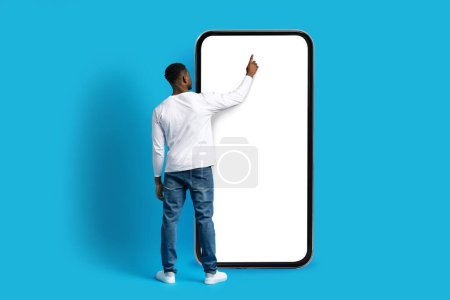 Téléchargez les photos : Homme afro-américain méconnaissable touchant écran blanc vierge d'un énorme téléphone, faisant des achats ou des opérations bancaires en ligne, à l'aide d'une application mobile, isolé sur fond bleu, maquette, espace de copie, pleine longueur - en image libre de droit