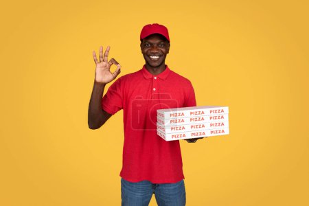Téléchargez les photos : Positif millénial afro-américain guy courier montrer de nombreuses boîtes à pizza, ok signe à la main, isolé sur fond orange. Annonce et offre, livraison de nourriture à domicile, bon service, approbation, recommandation - en image libre de droit