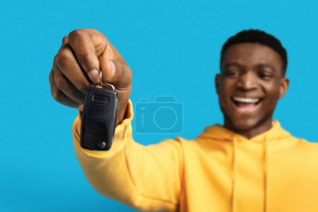 Foto de Llave automatizada de coche en mano de hombre negro, fondo estudio azul. Emocionado joven afroamericano feliz en sudadera con capucha amarilla que muestra la clave de su nuevo auto, celebrando la compra de automóviles - Imagen libre de derechos