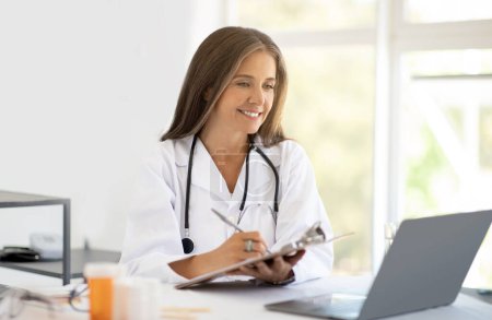 Photo for Cheerful adult caucasian nutritionist doctor in white coat at table makes notes in clipboard, looks at laptop in clinic office interior. Weight loss and health care, planning diet with professional - Royalty Free Image