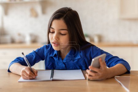 Photo for Arabic Schoolgirl Having Online Class Via Mobile Phone And Taking Notes Learning At Home. School Kid Girl Doing Homework Via Gadget, Writing Watching Distance Lesture. Modern Education Concept - Royalty Free Image