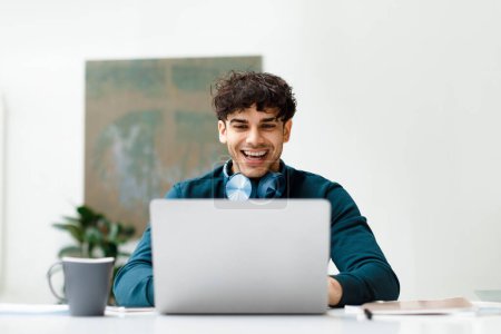 Téléchargez les photos : Homme freelance européen heureux travaillant sur ordinateur portable portant des écouteurs sur le cou et souriant, assis dans un bureau léger. Étudiant utilisant un ordinateur, regarder webinaire en ligne - en image libre de droit