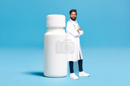 Photo for Indian man doctor in coat leaning at huge jar of medicines isolated on blue background, mockup space for your design. Pills for health care, medical ad and offer, pharmacy - Royalty Free Image
