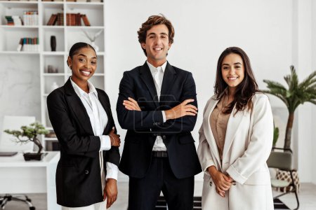 Photo for Three multiethnic businesspeople coworkers posing standing in modern office indoors, smiling at camera. Multiracial colleagues expressing confidence in career success - Royalty Free Image