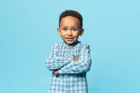 Foto de Retrato de niño afroamericano alegre posando ocasionalmente con los brazos cruzados sobre fondo azul, niño varón negro sonriendo y mirando a la cámara, espacio para copiar - Imagen libre de derechos