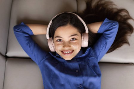 Téléchargez les photos : Profiter de la musique en ligne. Petite fille joyeuse écoutant de la musique portant des écouteurs sans fil allongés sur le canapé, se relaxant à la maison, vue d'en haut, souriant à la caméra. Concept amusant musical - en image libre de droit