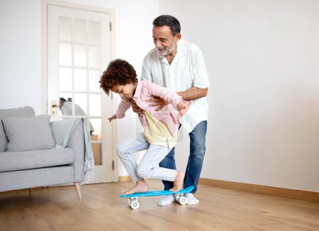 Photo for Smiling Latin Grandfather Guides Grandsons First Skateboard Ride at Home, Holding Kid Boy and Assisting Him on Board In Modern Living Room, Enjoying Quality Time and Fun Together - Royalty Free Image