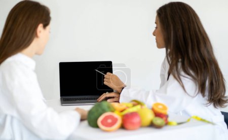 Photo for Adult caucasian nutritionist doctor in white coat shows weight loss plan on laptop blank screen to young woman patient in clinic office interior. Online recommendation by professional, health care - Royalty Free Image