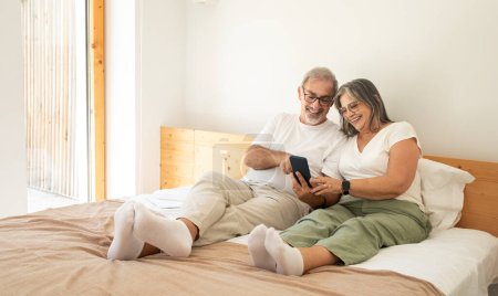 Photo for Cheerful old caucasian husband and wife lie in bed, chatting on phone, enjoy good morning together in bedroom interior. Weekend with app, gadget, relationships and love, lifestyle - Royalty Free Image