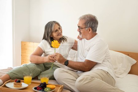 Photo for Happy old caucasian man give flower to wife on bed, have romantic breakfast, enjoy good morning together in bedroom interior. Rest at weekend, relationships and love at free time - Royalty Free Image