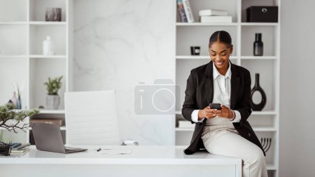 Téléchargez les photos : Femme d'affaires noire réussie en tenue formelle assise au bureau avec ordinateur portable et utilisant un téléphone portable bavardant avec le client ou vérifiant les e-mails, travaillant au bureau, panorama - en image libre de droit