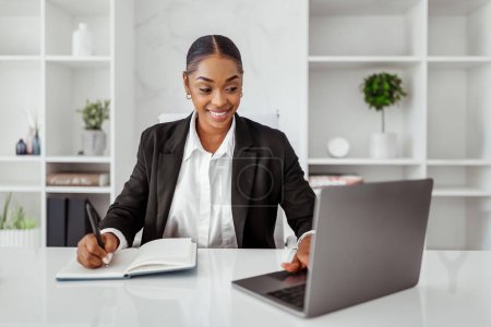 Téléchargez les photos : Joyeux jeune femme noire entrepreneure assister à une réunion d'affaires en ligne, dame assise devant un ordinateur portable et prenant des notes à l'intérieur du bureau léger, espace de copie - en image libre de droit