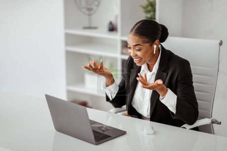 Téléchargez les photos : Femme d'affaires afro-américaine souriante en tenue formelle ayant un appel vidéo sur ordinateur portable, portant des écouteurs et gestualisant à la webcaméra, travaillant à l'intérieur du bureau. Communication commerciale à distance - en image libre de droit