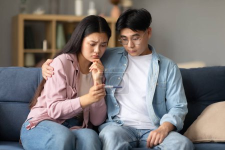 Photo for Infertility. Unhappy Japanese Couple Holding Negative Pregnancy Test Result Sitting On Sofa, Hugging And Supporting Each Other In Problem At Home. Reproduction, Childbirth Healthcare Struggle - Royalty Free Image