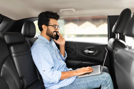 Photo for Positive young indian man freelancer working online while sitting at backseat in taxi, eastern guy entrepreneur typing on laptop with blank screen and having phone conversation, copy space, side view - Royalty Free Image