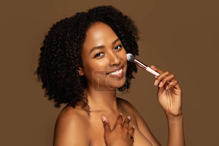 Photo for Closeup of cheerful pretty half-naked young black woman with bushy hair holding makeup brush, isolated on brown background. African american lady applying blush or foundation on her face - Royalty Free Image
