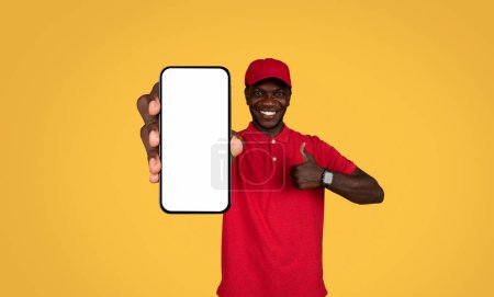 Photo for Cheerful handsome millennial african american guy courier in uniform show phone with blank screen and thumb up, isolated on orange studio background. Great service, approving app delivery - Royalty Free Image
