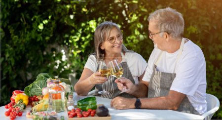 Photo for Smiling old caucasian husband and wife in aprons cheers glasses of wine at table in home garden, outdoor. Romantic date, dinner and relationship, love and food, holiday celebration - Royalty Free Image