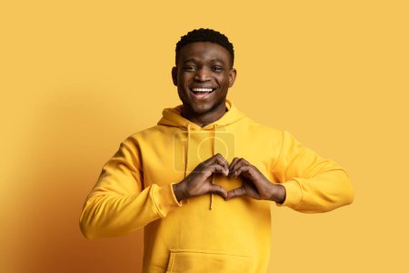 Photo for Cheerful handsome millennial black guy wearing yellow hoodie holding hands on chest in heart-shaped symbol and smiling, expressing love isolated on yellow background, copy space - Royalty Free Image