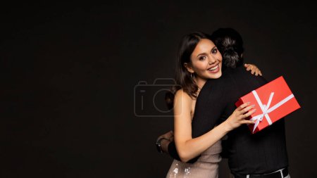 Foto de Sonriente joven atractiva sosteniendo regalo de Navidad caja roja, abrazando a su novio. Pareja cariñosa celebrando Año Nuevo 2024 juntos sobre fondo negro, espacio para copiar, pancarta - Imagen libre de derechos