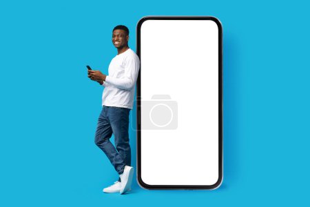 Photo for Nice mobile app. Cheerful young black guy in casual outfit standing next to big phone with white blank screen, using smartphone and smiling, mockup, copy space, isolated on blue background - Royalty Free Image