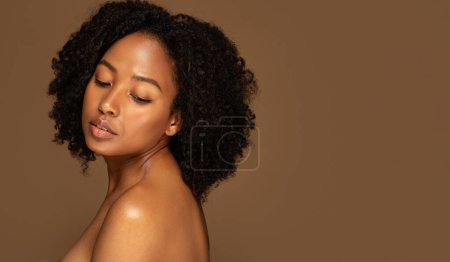 Foto de Señora afroamericana joven desnuda posando sobre fondo de estudio marrón, vista lateral de la mujer negra milenaria en topless mirando hacia abajo, espacio para copiar, panorama. Concepto de sensualidad femenina - Imagen libre de derechos