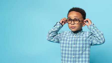 Photo for African american little boy wearing eyeglasses having poor eyesight posing standing on blue studio background, panorama with copy space. Kids eyes health - Royalty Free Image