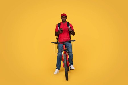 Photo for Smiling millennial african american guy courier in uniform with backpack on bike, point finger at smartphone, isolated on orange studio background. Recommendation app, gadget for delivery, work - Royalty Free Image