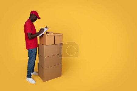 Photo for Cheerful millennial african american guy courier in uniform making notes in warehouse, cheks many boxes, isolated on orange studio background. Ad and offer, delivery, work and service - Royalty Free Image
