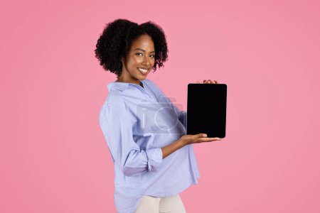Foto de Positivo joven embarazada negro en la tableta show casual con pantalla vacía, recomienda aplicación, aislado en el fondo del estudio de color rosa. Dispositivo para esperar bebé, chat y redes sociales - Imagen libre de derechos